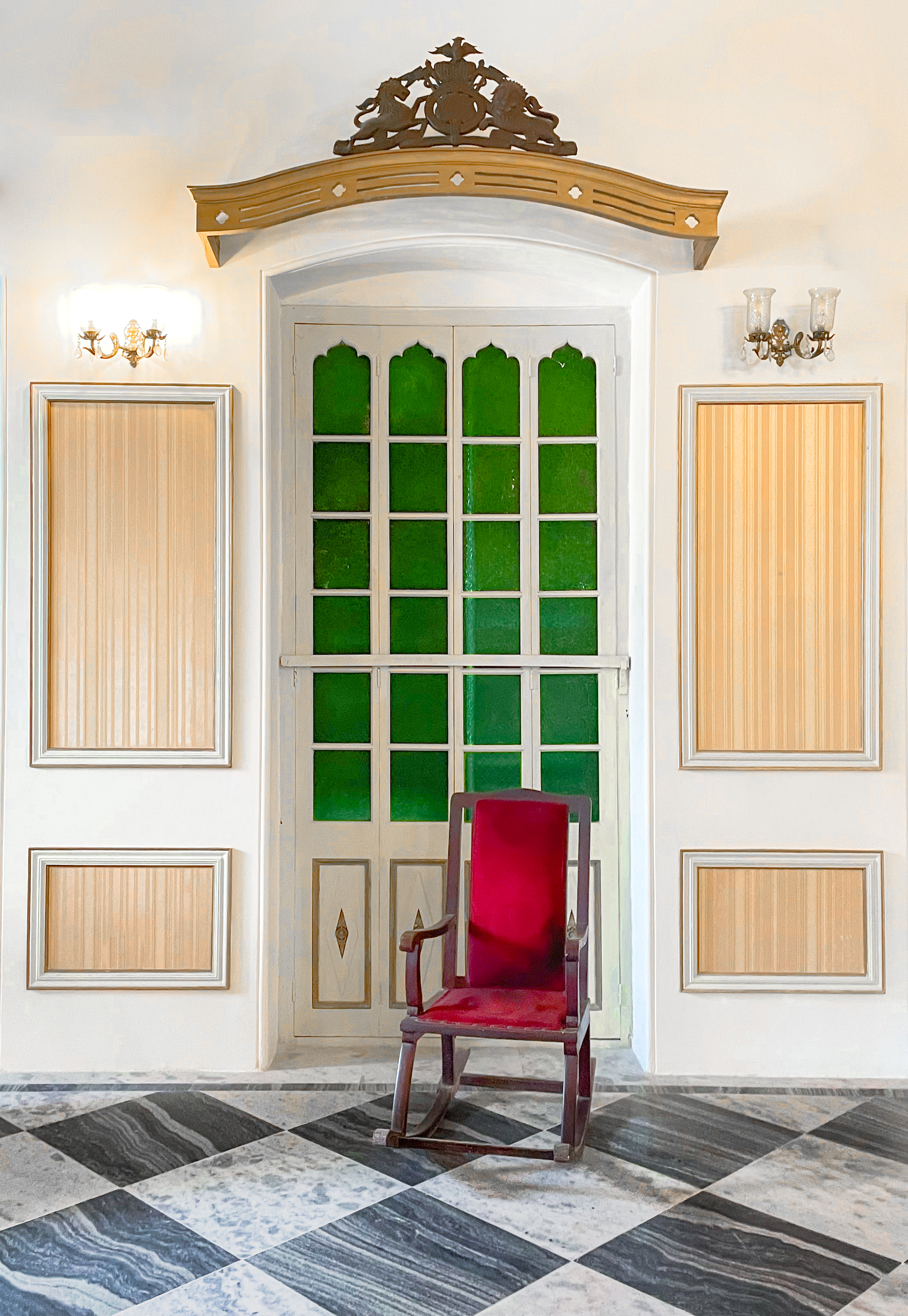 Framed by green stained glass and golden paneling, this corner of the palace reflects the quiet aesthetic of Mughal design. The black-and-white marble floor and carved pediment create a symmetry of pattern and form. A lone velvet rocking chair completes the scene.