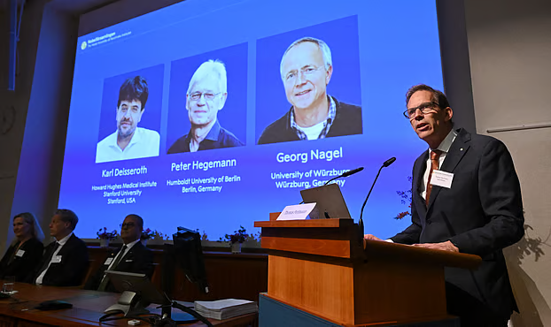 Nobel Committee Secretary General Thomas Perlmann (R) speaks to the media in front of a screen displaying the names of this year’s laureates (L-R) Karl Deisseroth, Peter Hegemann and Georg Nagel during the announcement of the winners of the 2026 Nobel Prize in Physiology or Medicine at the Karolinska Institute in Stockholm, Sweden, on 5 October 2026.AFP